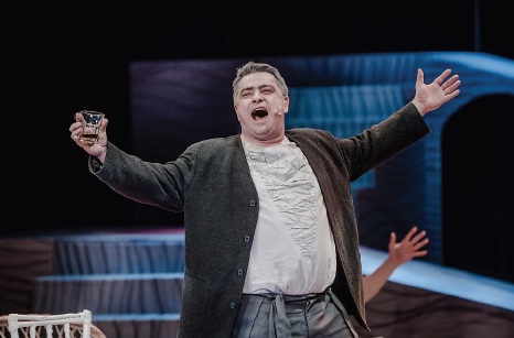 Tomasz Lorek jest śpiewakiem w średnim wieku, występuje w białej koszuli i brązowym surducie, ręce ma szeroko rozłożone